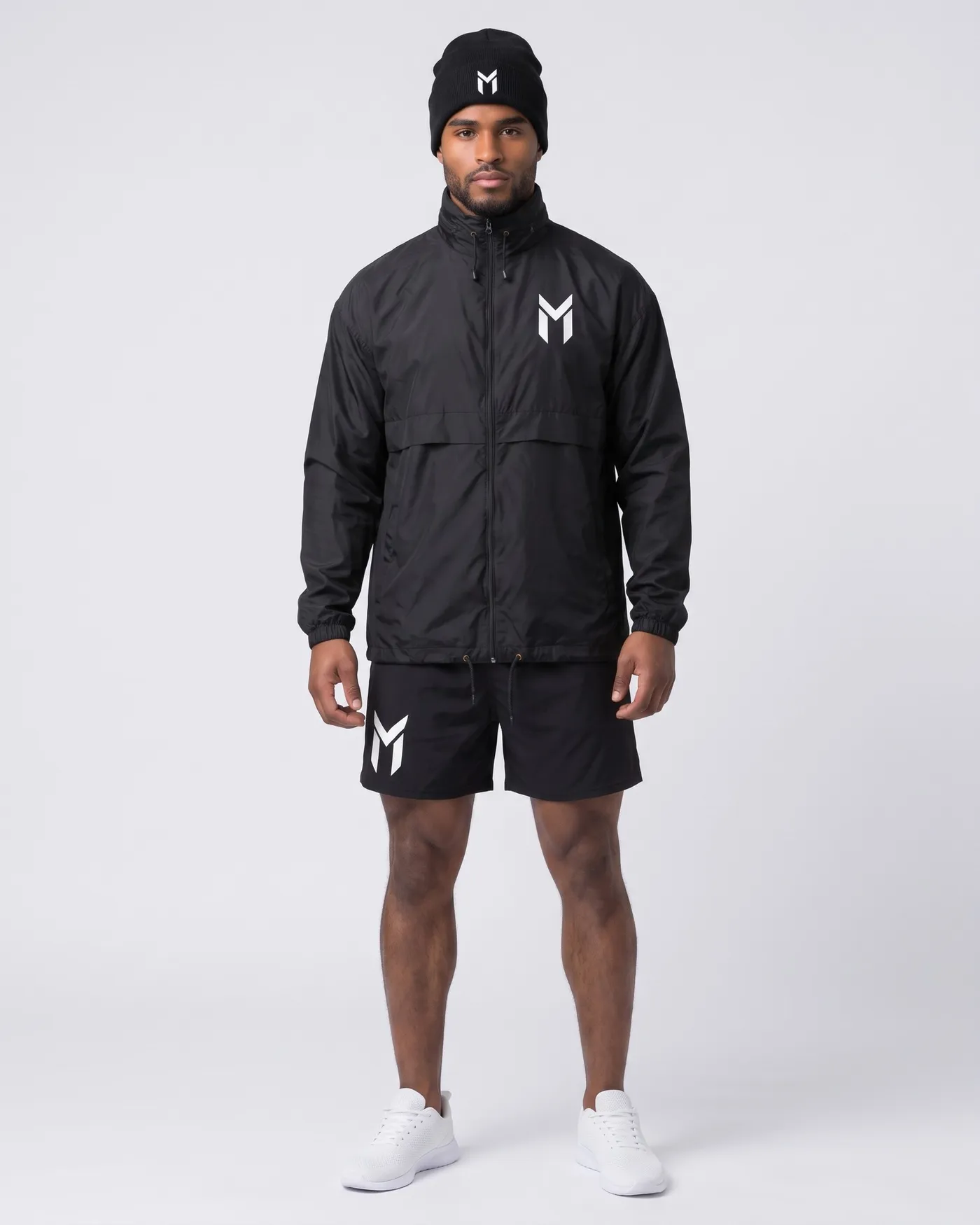 Person in schwarzem Windbreaker, schwarzen Gym Shorts und schwarzer Beanie vor hellgrauem Grund